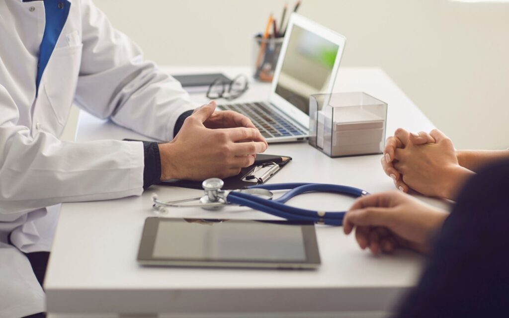 medico_pacientes Médico em jaleco branco e estetoscópio conversando com paciente em consultório, simbolizando a importância do acompanhamento profissional de saúde.